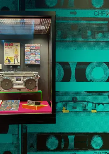 A museum display featuring a vintage boombox, hip-hop event posters, cassette tapes, and a turquoise background with a cassette tape graphic.